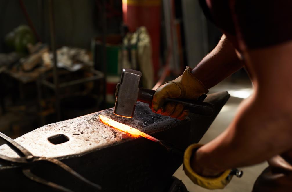 Close-up of unrecognizable blacksmith standing at steel anvil and shaping heated metal bar with hammer in workshop