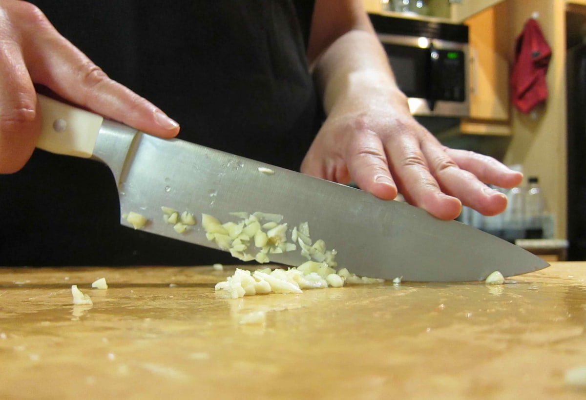 A chef knife rocking through garlic on a wooden board